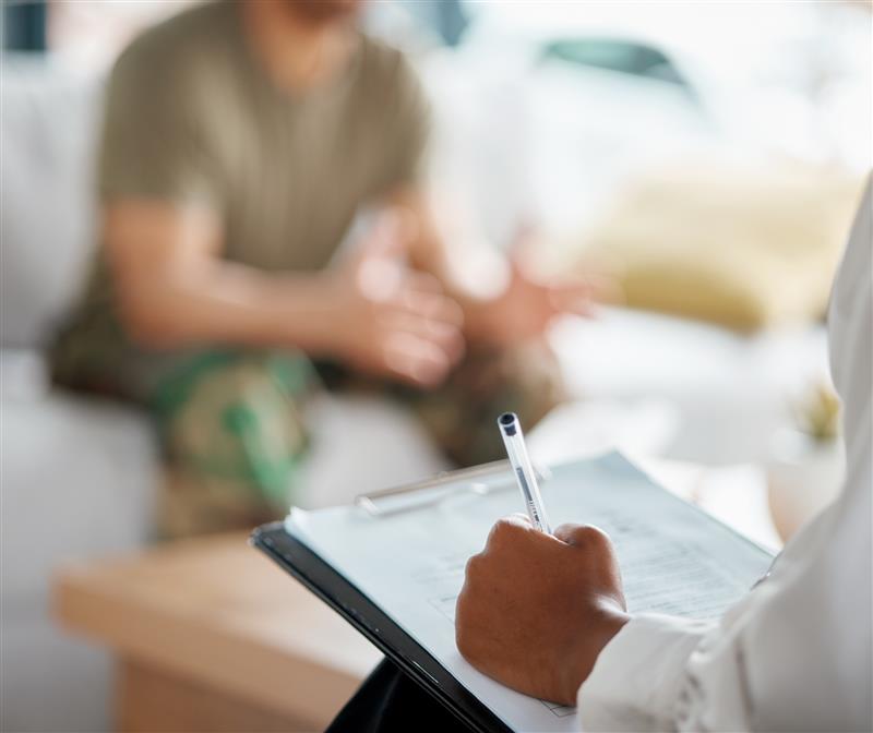 Mostly-blurred image with two people, one sitting and talking to the other writing notes about their conversation.